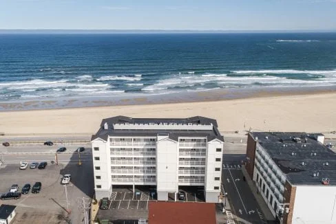 Aerial view of a beachfront building facing the ocean, with a sandy beach and parked cars visible in the foreground. Waves can be seen crashing in the distance.