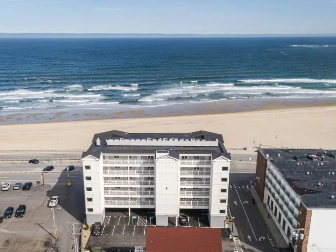 Aerial view of a beachfront building facing the ocean, with a sandy beach and parked cars visible in the foreground. Waves can be seen crashing in the distance.