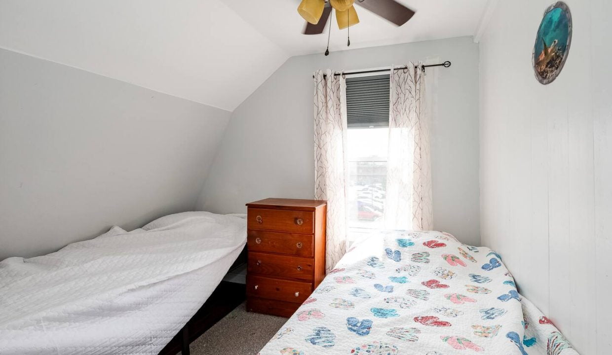 A small, brightly-lit attic bedroom with two single beds, a wooden dresser, and a ceiling fan.