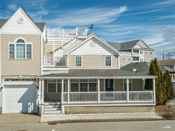 Two-story beige house with white trim, a large wraparound porch, attached garage, and rooftop deck, situated on a paved street under a partly cloudy sky.