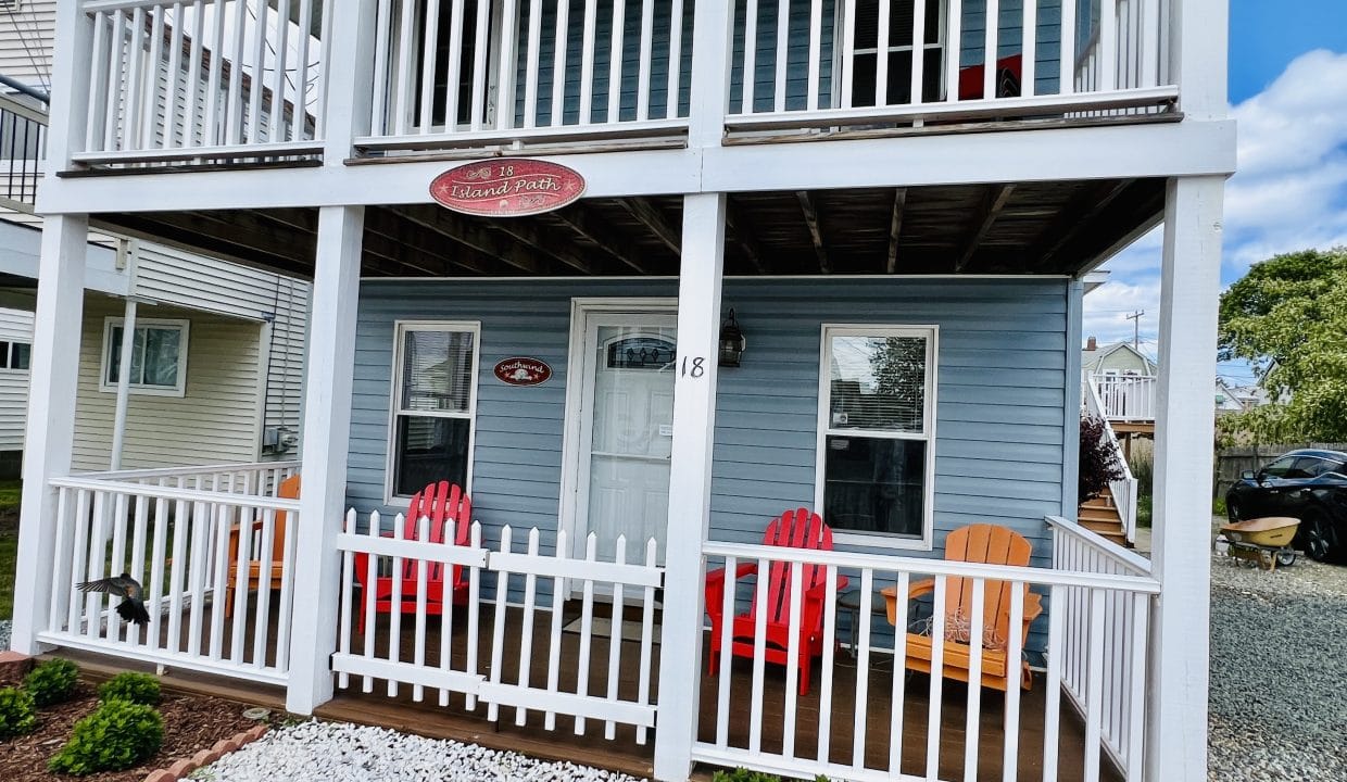 A two-story blue house with a white railing porch, four colorful Adirondack chairs, a stone garden in front, and a sign above the door.