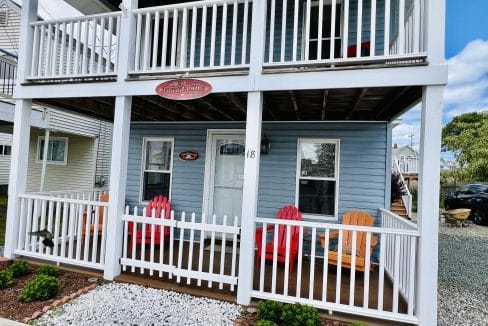 A two-story blue house with a white railing porch, four colorful Adirondack chairs, a stone garden in front, and a sign above the door.