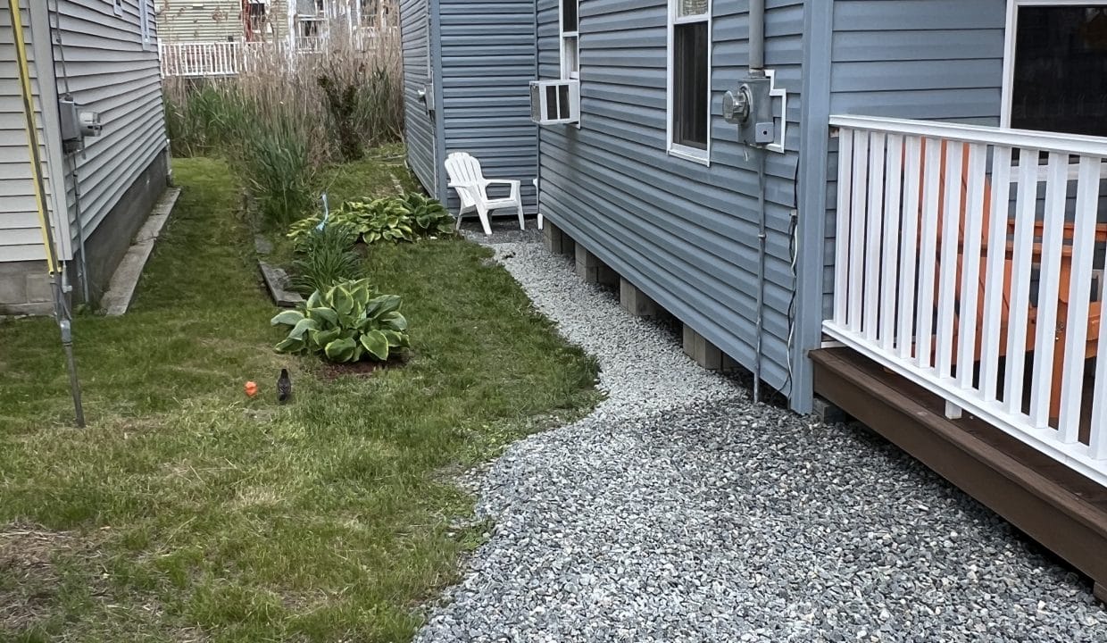 A narrow yard beside a gray house features a patch of gravel, a small green plant, a white plastic chair, and grass along the side of another house.