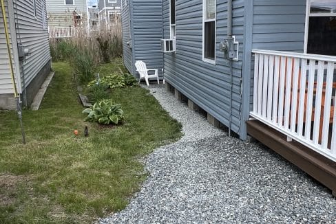 A narrow yard beside a gray house features a patch of gravel, a small green plant, a white plastic chair, and grass along the side of another house.