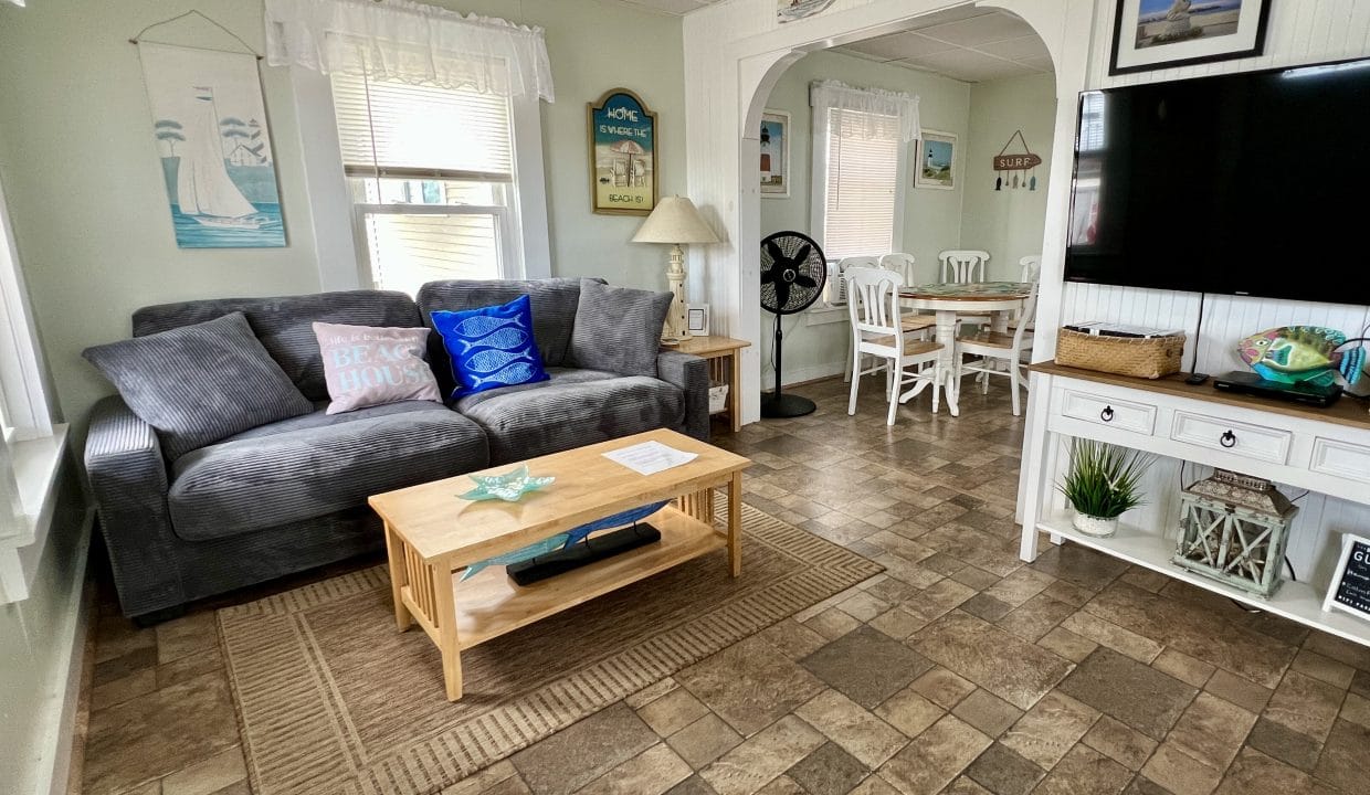 Living room with a gray sofa, coffee table, TV on a white stand, nautical decor, and an adjacent dining area with a white table and chairs.
