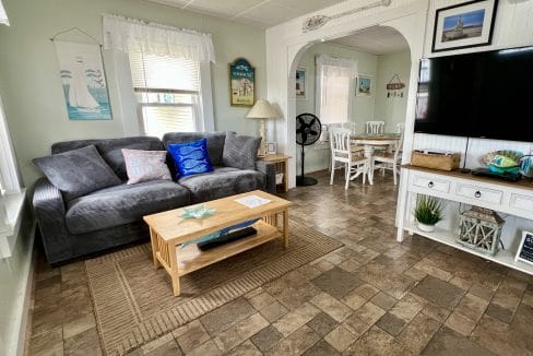 Living room with a gray sofa, coffee table, TV on a white stand, nautical decor, and an adjacent dining area with a white table and chairs.