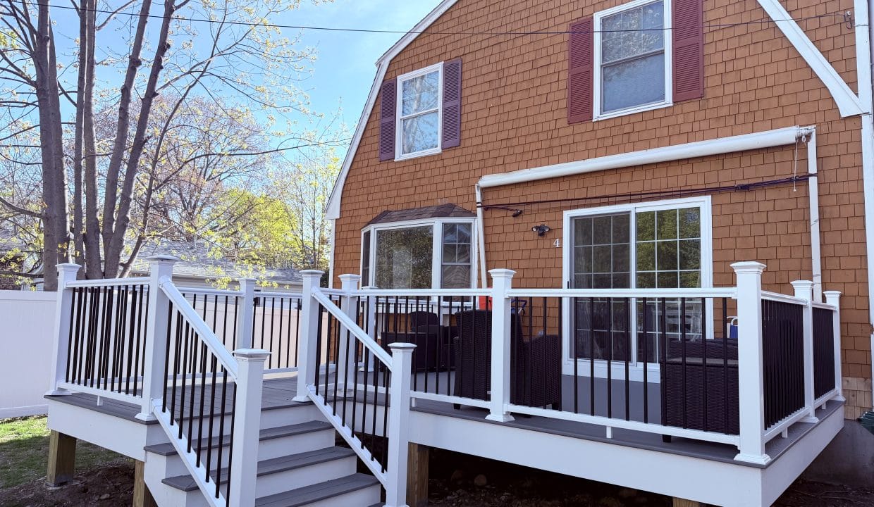 A brown house with white trim features a raised deck with white railings and black balusters, patio furniture, and stairs leading to the backyard.