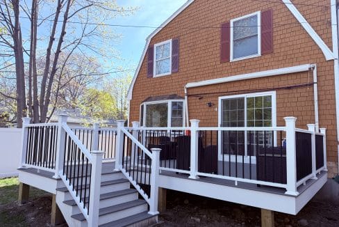 A brown house with white trim features a raised deck with white railings and black balusters, patio furniture, and stairs leading to the backyard.