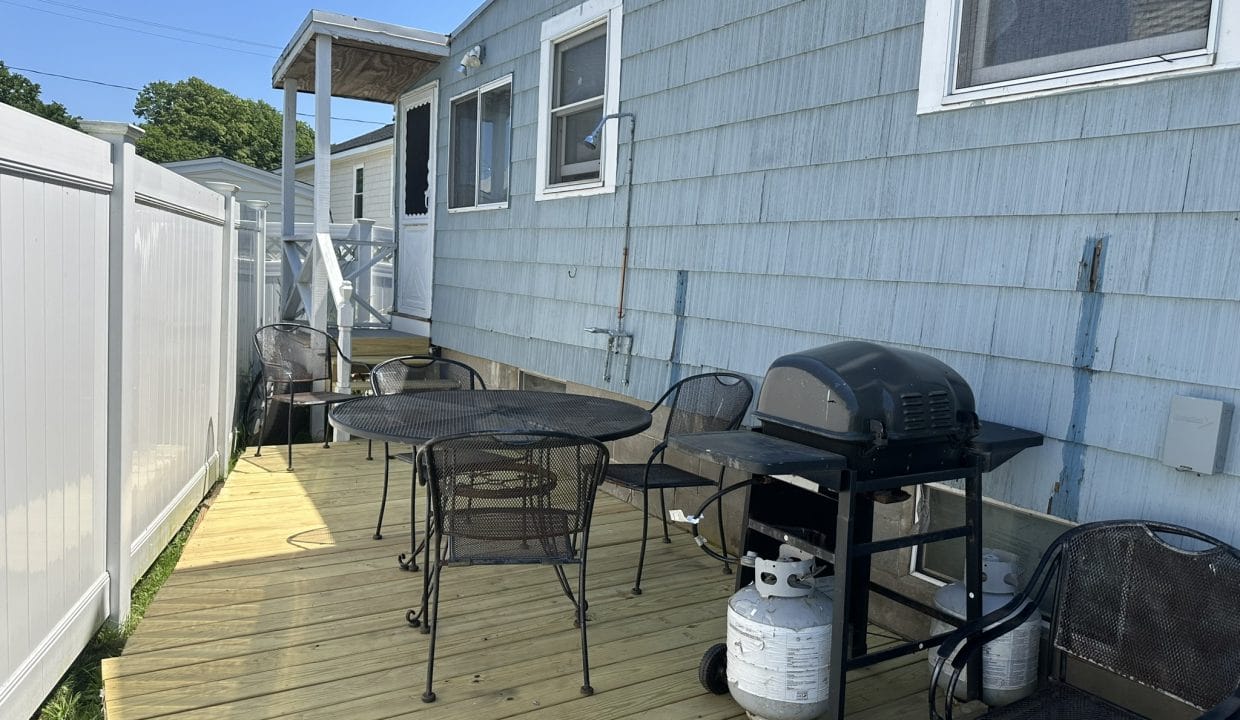 A wooden deck with a round table, several chairs, and a gas grill sits next to a light blue house with white windows and a fence.