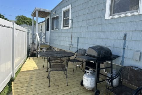 A wooden deck with a round table, several chairs, and a gas grill sits next to a light blue house with white windows and a fence.