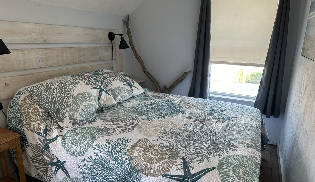 A neatly made bed with a seashell and starfish-patterned quilt is placed by a window with dark curtains and a beige blind in a small, simply decorated bedroom.