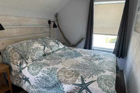 A neatly made bed with a seashell and starfish-patterned quilt is placed by a window with dark curtains and a beige blind in a small, simply decorated bedroom.