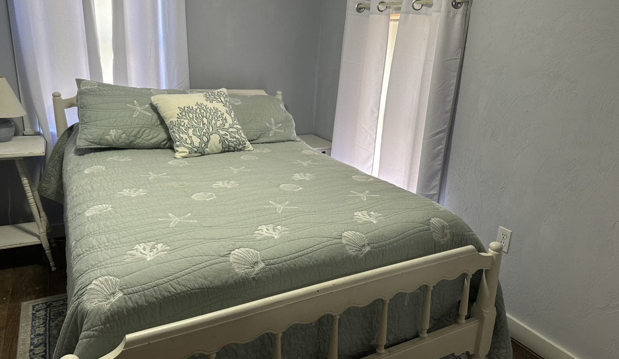 A neatly made bed with sea-themed bedding and pillows is positioned near two curtained windows in a small, light-gray bedroom. A nightstand with a lamp sits beside the bed.