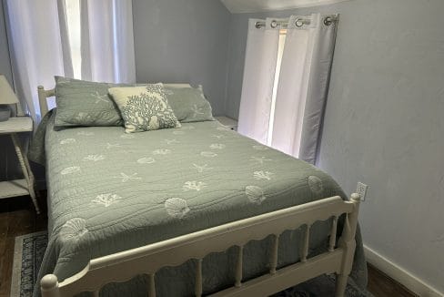 A neatly made bed with sea-themed bedding and pillows is positioned near two curtained windows in a small, light-gray bedroom. A nightstand with a lamp sits beside the bed.