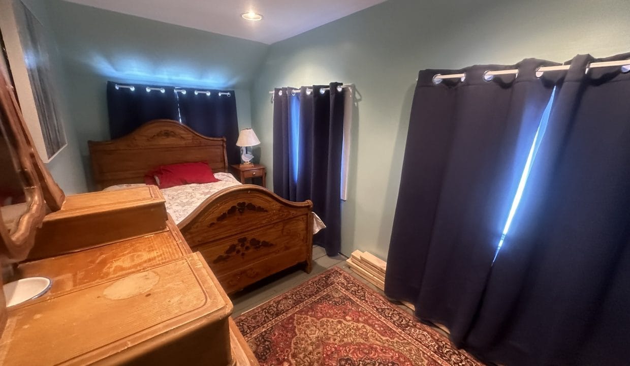 A small bedroom with a wooden bed, matching dresser, red and tan rug, nightstand with a lamp, and windows covered by dark curtains.