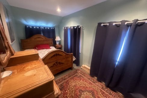 A small bedroom with a wooden bed, matching dresser, red and tan rug, nightstand with a lamp, and windows covered by dark curtains.