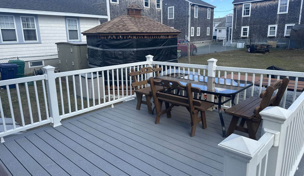 A residential backyard featuring a wooden deck with a table and chairs, bordered by a white railing, with a gazebo and houses in the background.