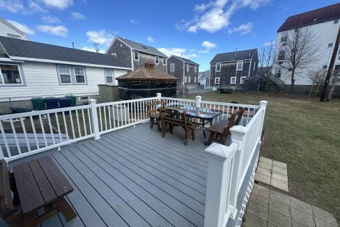 A residential backyard with a wooden deck, patio furniture, and houses in the background under a clear blue sky.