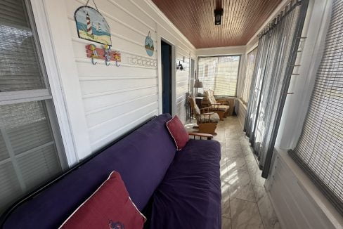A cozy, sunlit enclosed porch with a purple couch, decorative pillows, a rocking chair, and nautical-themed wall art.