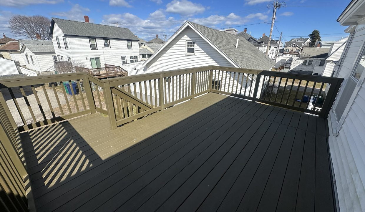 A residential outdoor wooden deck with dark flooring, surrounded by a light wooden railing, overlooking a neighborhood with houses and clear blue skies.