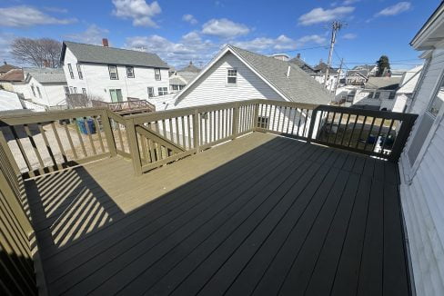 A residential outdoor wooden deck with dark flooring, surrounded by a light wooden railing, overlooking a neighborhood with houses and clear blue skies.