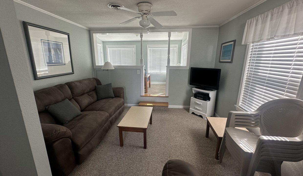 A neatly arranged living room with a brown sofa, wooden coffee table, television, and white chairs, featuring ample natural light from windows with blinds.