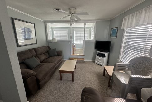 A neatly arranged living room with a brown sofa, wooden coffee table, television, and white chairs, featuring ample natural light from windows with blinds.