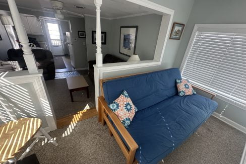 A bright, sunlit living room with a blue futon sofa and decorative pillows, overlooking an interior space through a square archway.