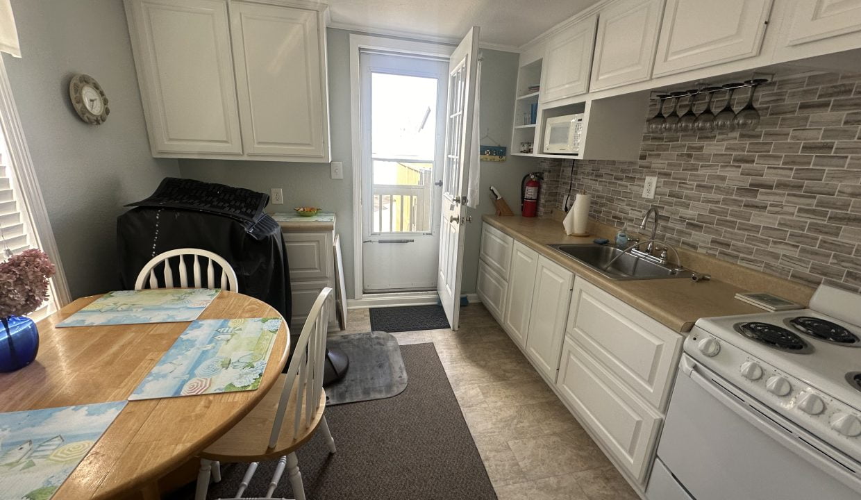 A bright, compact kitchen with white cabinetry, modern appliances, and a wooden dining table set near the entrance to a balcony.