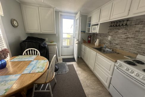 A bright, compact kitchen with white cabinetry, modern appliances, and a wooden dining table set near the entrance to a balcony.