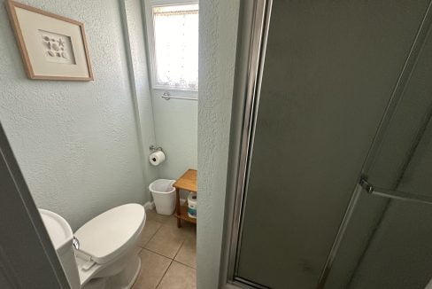 Small bathroom with a toilet, corner shower, and a window with blinds.