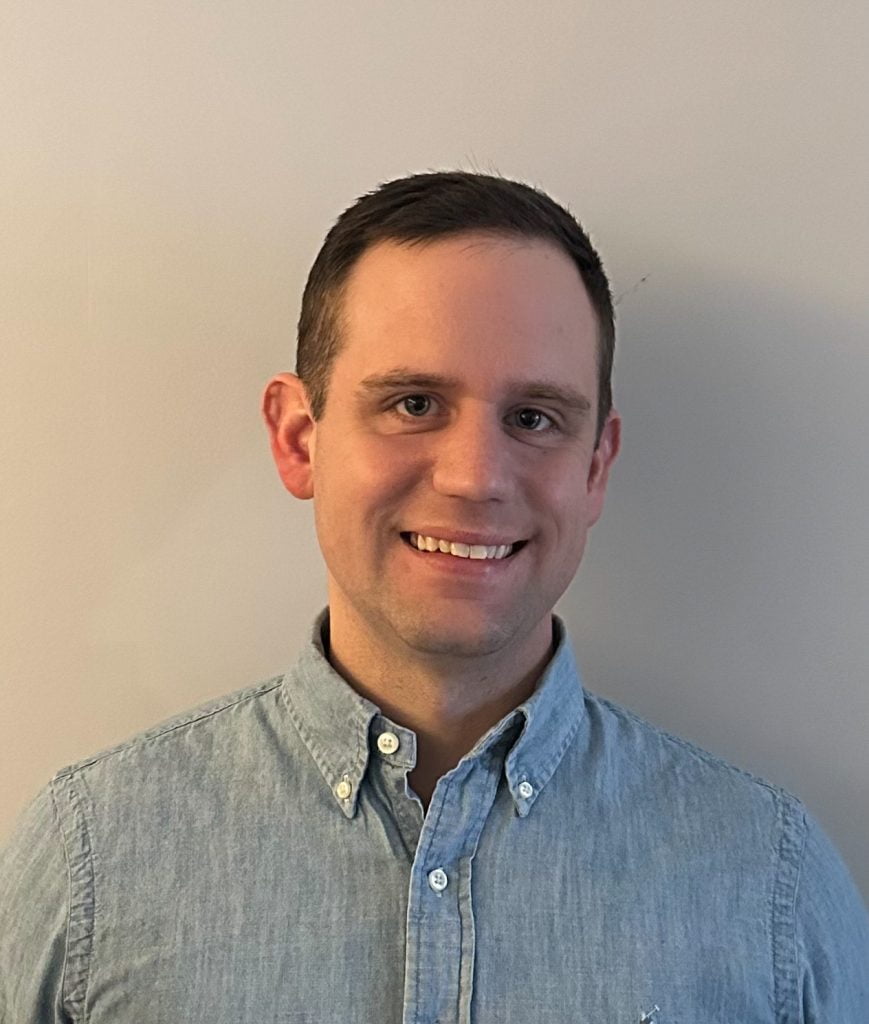 A man in a blue shirt smiling in front of a white wall.