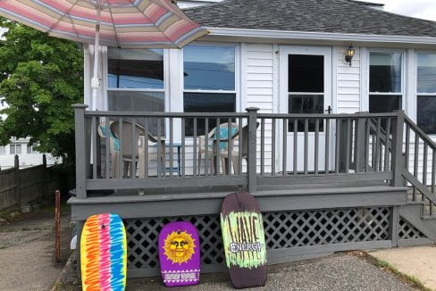 A wooden deck with a patio table and umbrella outside a house, accompanied by three colorful bodyboards leaning against the railing.