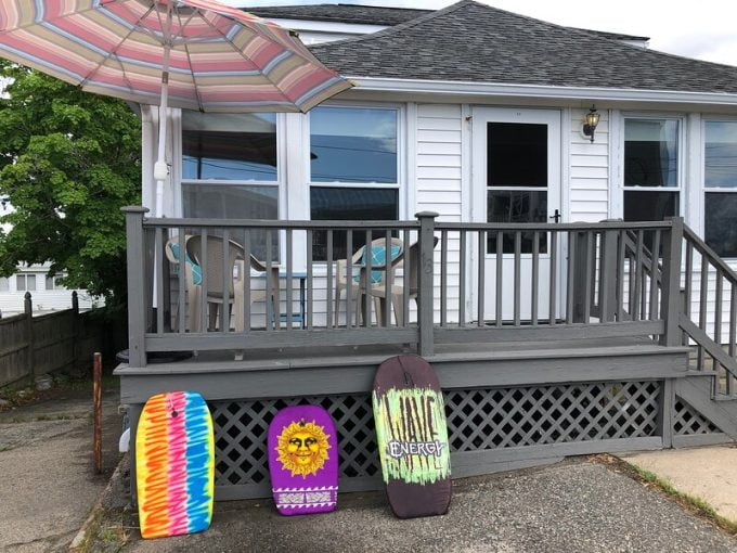 A wooden deck with a patio table and umbrella outside a house, accompanied by three colorful bodyboards leaning against the railing.