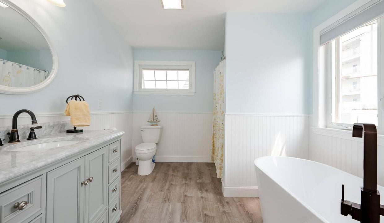 A bright, modern bathroom with blue walls, white wainscoting, and wood-like flooring, featuring a freestanding tub, vanity with sink, and toilet.