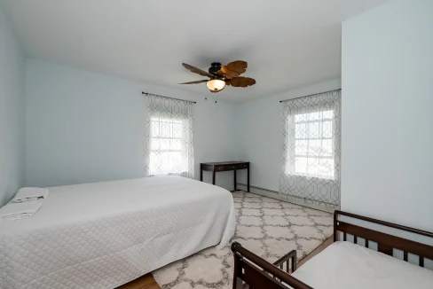 A bright, minimally furnished bedroom with a double bed, a ceiling fan, and two windows with patterned curtains.