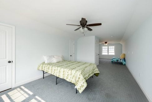 A bright, sparsely furnished bedroom with a single bed, ceiling fan, and a door leading to another room.