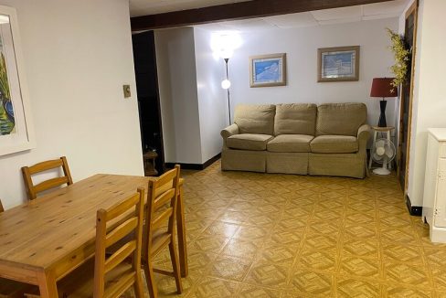 A cozy living space with a wooden dining table and a beige sofa, featuring yellow-patterned linoleum flooring and wall-mounted artwork.