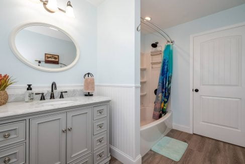 A well-lit bathroom with a gray vanity, round mirror, and a shower with a colorful curtain.
