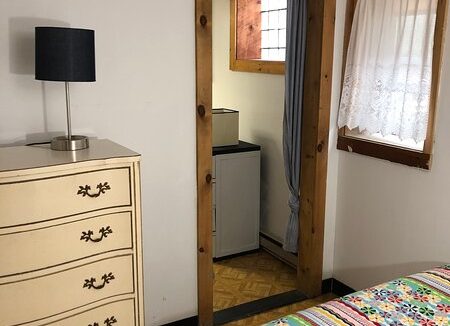 A modestly furnished room with a patterned bedspread, a wooden dresser, a lamp, and a view into an adjacent room with a refrigerator.
