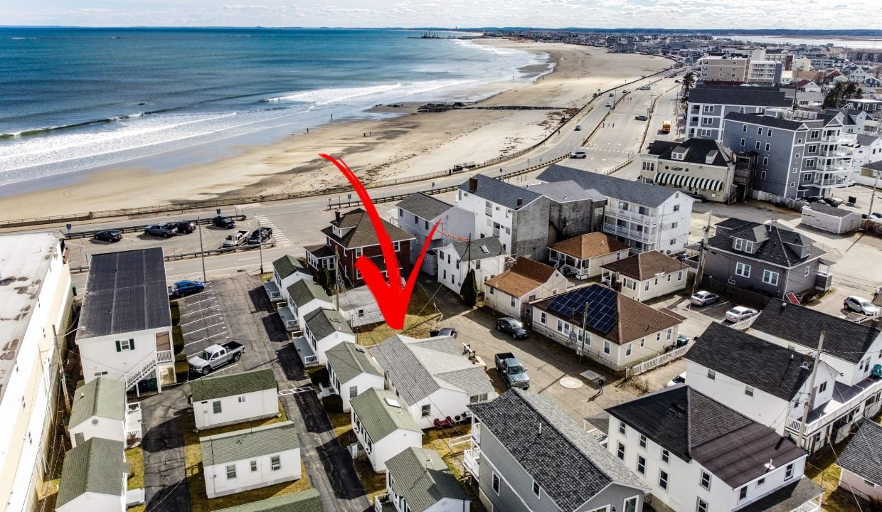Aerial view of a coastal town with buildings near a sandy beach, indicated by a red arrow.