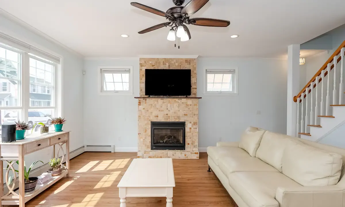 Bright living room with a stone fireplace, wall-mounted tv, ceiling fan, and light-colored furniture.