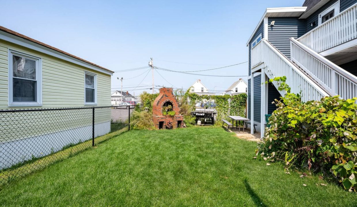 Backyard with a well-maintained lawn, a brick barbecue grill, surrounded by a chain-link fence, and flanked by two houses.