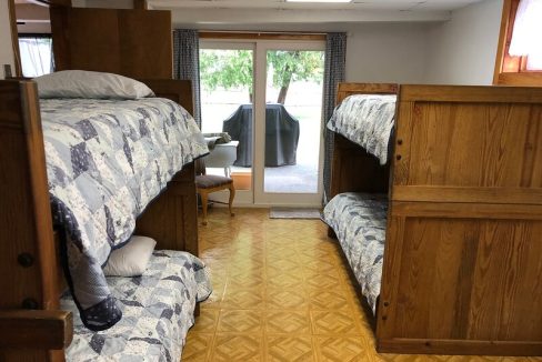 A dormitory room with two wooden bunk beds, patterned bedding, and a desk near the window.