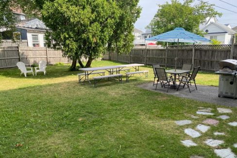 A backyard with picnic tables, chairs, a green lawn, and a swimming pool in the distance.