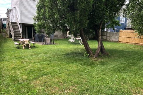 Backyard with a large tree, lawn, two picnic tables, and a grill next to a white house with stairs.