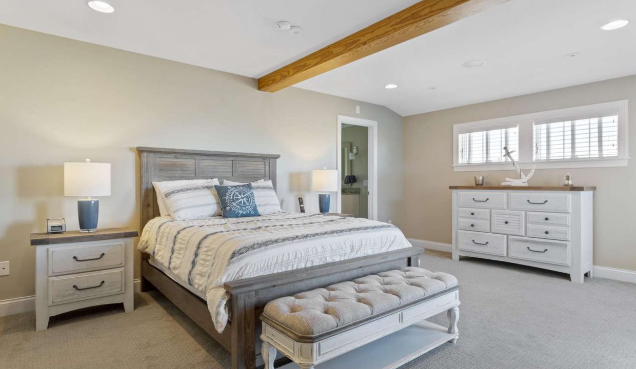 Spacious bedroom with neutral colors, featuring a king-sized bed, wooden ceiling beam, and a dresser.