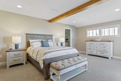 Spacious bedroom with neutral colors, featuring a king-sized bed, wooden ceiling beam, and a dresser.