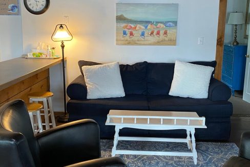 A cozy living room with a dark blue sofa, white pillows, a small white coffee table, and a beach painting on the wall.