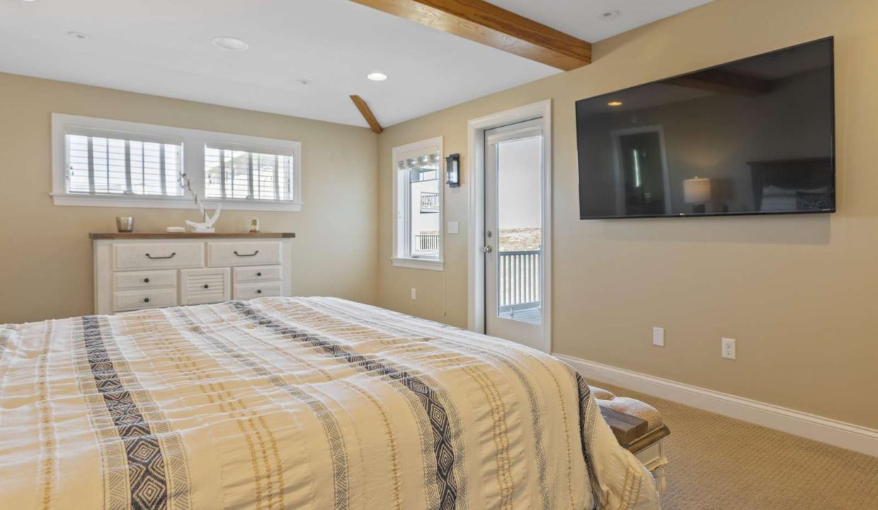 A bright and cozy bedroom featuring a neatly made bed, a dresser under a window, exposed ceiling beams, and a wall-mounted flat-screen tv.